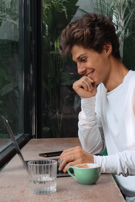 A young man with short brown hair smiling at a laptop in a café with a cup of coffee.